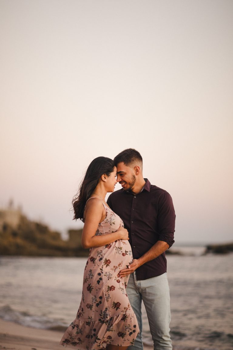 casal na praia, mamã grávida, de frente um para o outro, com testas encostadas. ela de vestido claro com rosas pequeninas, de alças e o papá de camisa roxa e calças de ganga.