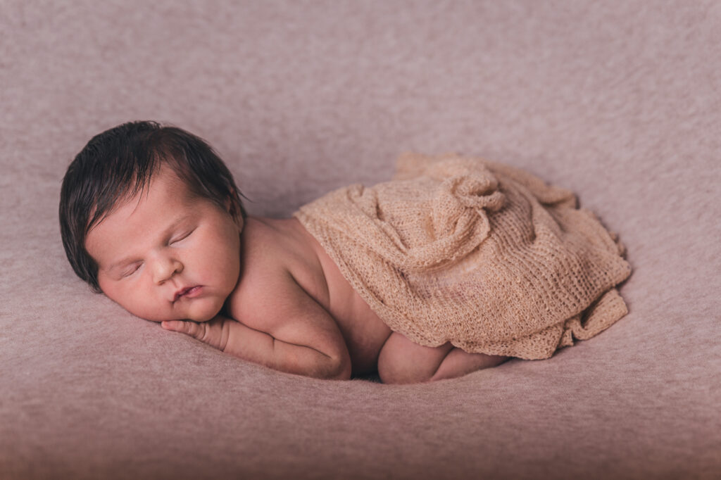 Fotografia de bebé recém-nascido a dormir enrolado num pano bege, deitado de lado sobre fundo rosado.