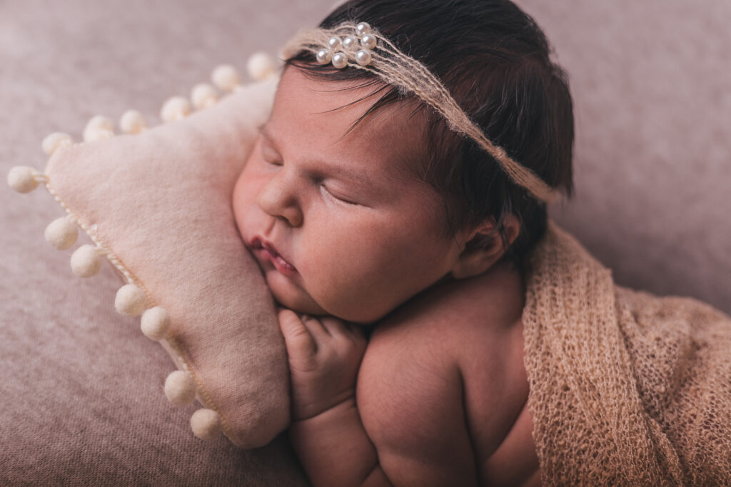 Bebé recém-nascido com faixa de pérolas na cabeça, a dormir com a bochecha apoiada numa mini almofada decorada.