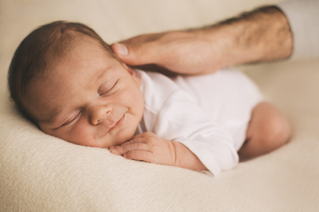 Bebé recém-nascido com leve sorriso a dormir sereno, de branco, enquanto é acariciado pelo pai.