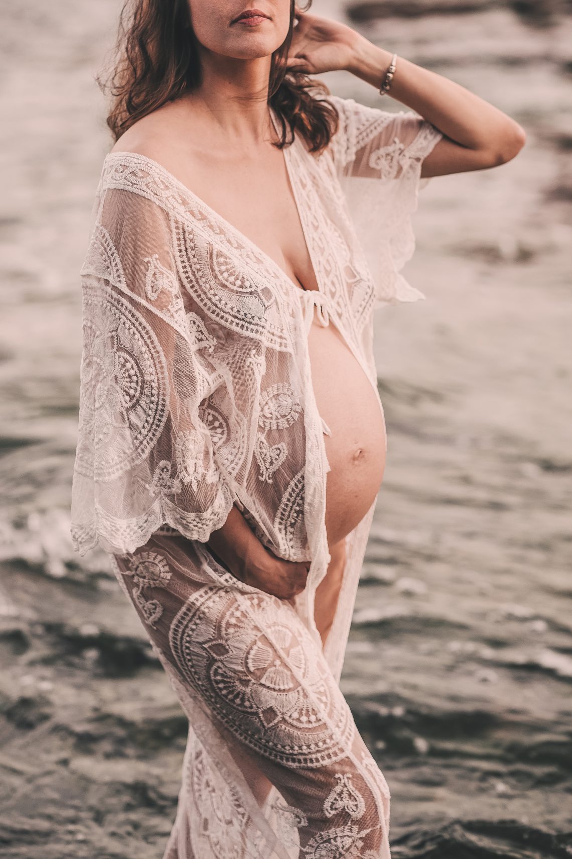mulher gravida de frente, com o mar de fundo, com ondas. mulher veste um kimono branco de rendas, tem a barriga a mostra, e passa. mão pelo cabelo. mamã, maternidade real, usando roupa para uma sessão de grávida