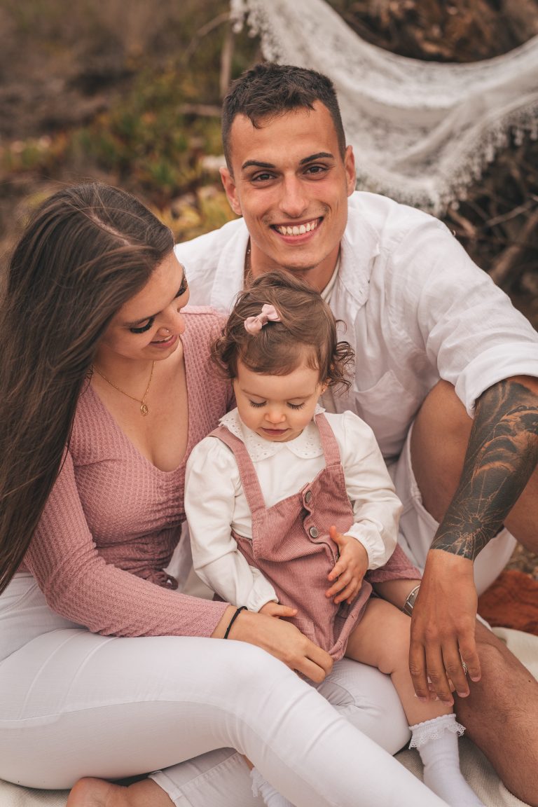 casal e sua filha de 2 anos sentados no campo. o pai olha de frene para a fotografia, a mãe tem a sua pequena ao colo e olha para ela, enquanto ela brinca.
