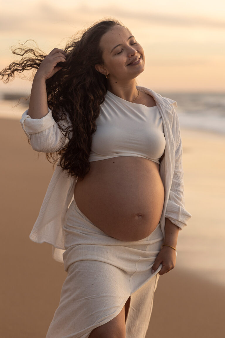 gravida na praia, ar sereno, uma mão a segurar o cabelo e outra agarrada a saia. tem a barriga a mostra e um top.